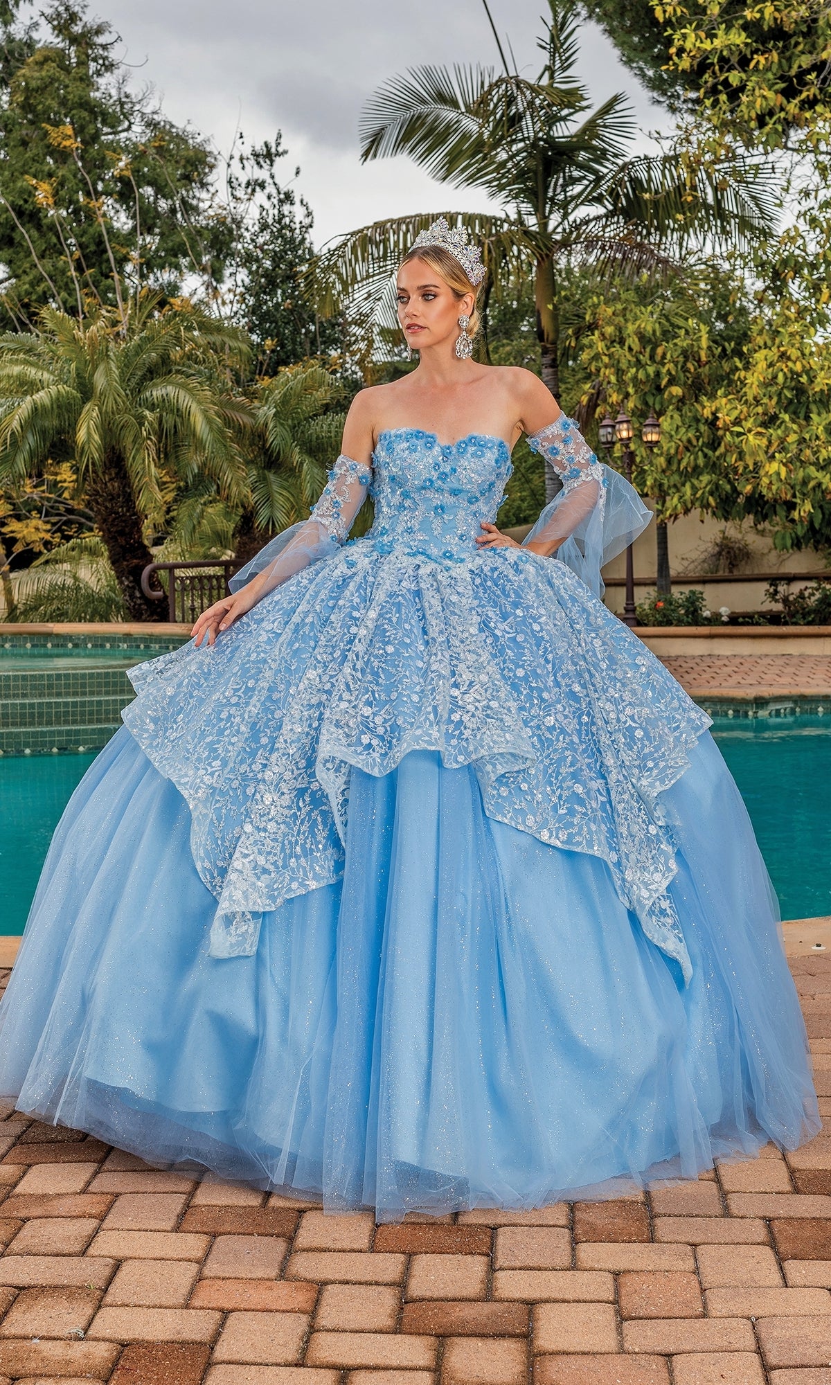 By the pool, a woman wearing the Quinceanera Dress 1773 By Dancing Queen—with lace, floral details, removable sheer sleeves, and a tiara—stands surrounded by lush greenery.
